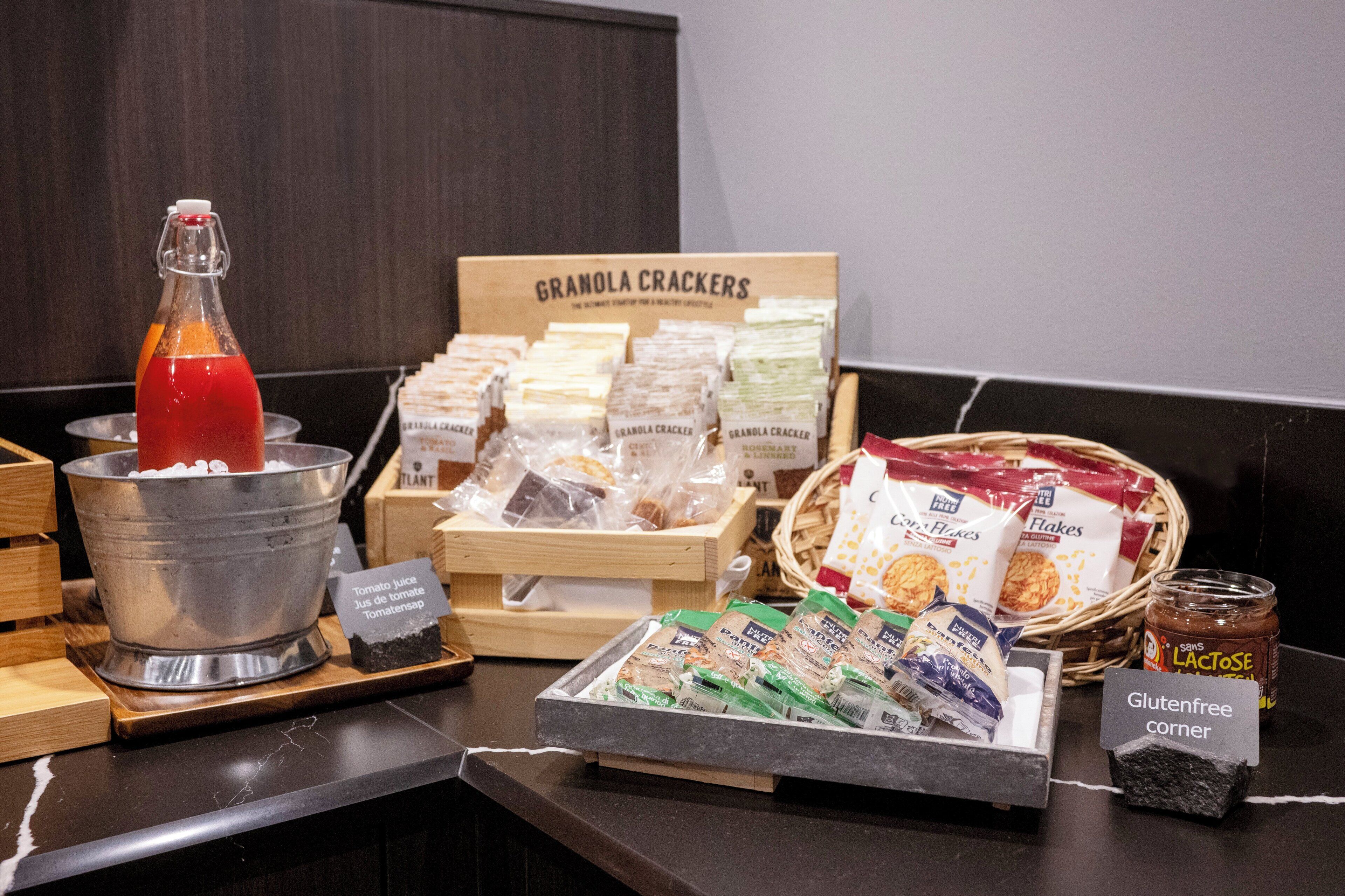 A breakfast buffet with granola crackers, tomato juice, lactose-free yogurt, gluten-free corner, and various cereal packets.
