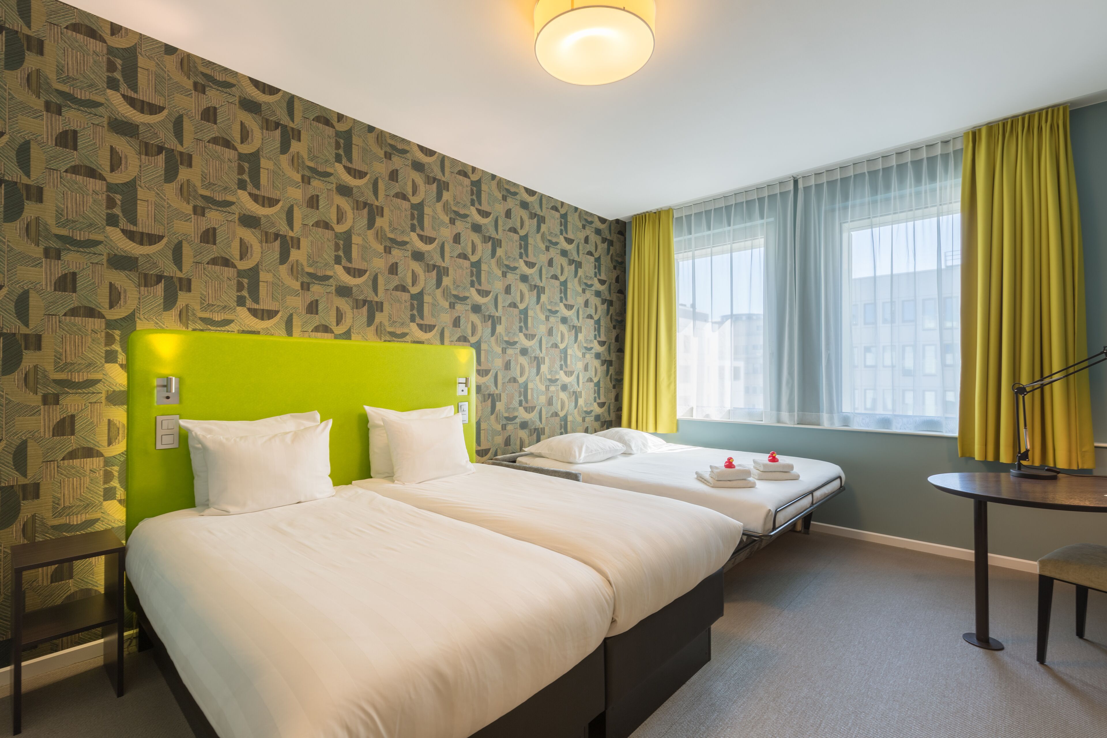 A hotel room with two beds, a green headboard, a small table, and a window with yellow curtains.