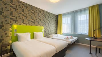 A hotel room with two beds, a green headboard, a small table, and a window with yellow curtains.