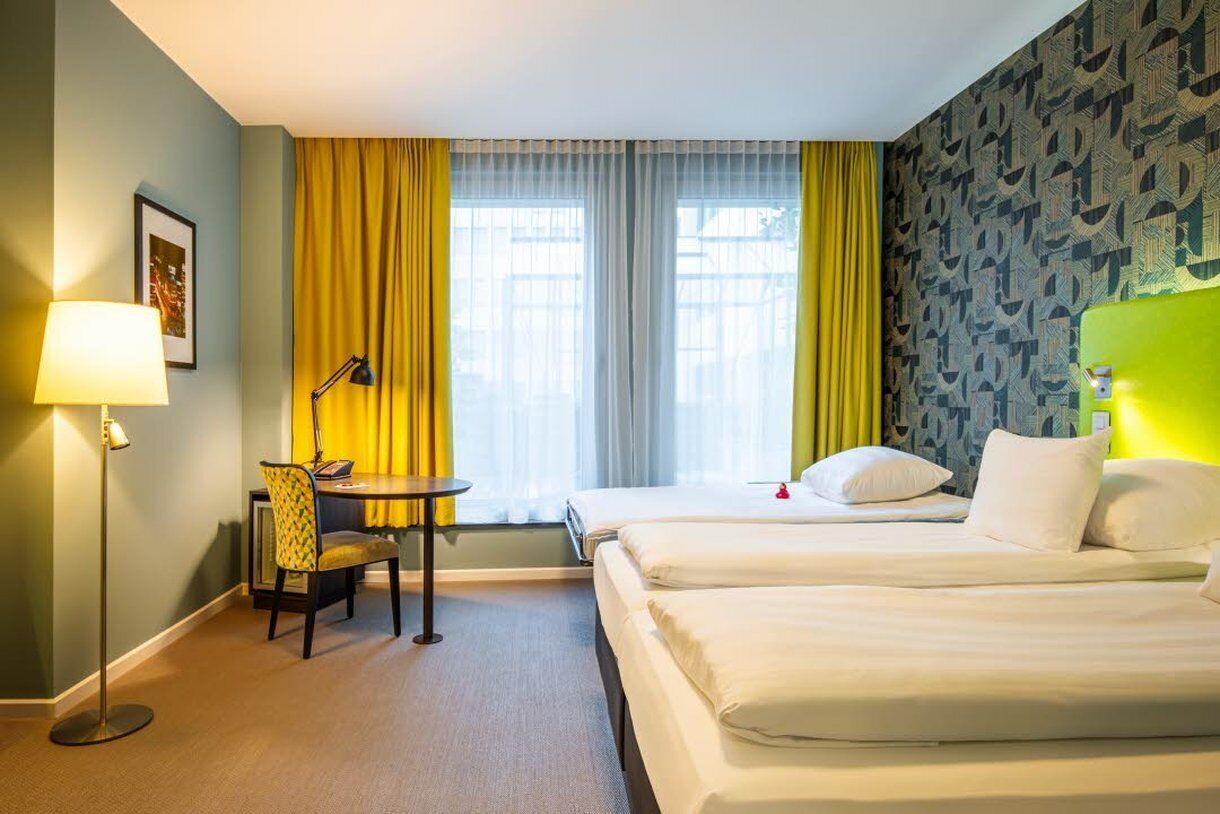 A hotel room with two beds, a desk with a chair, a lamp, and a window with yellow curtains.