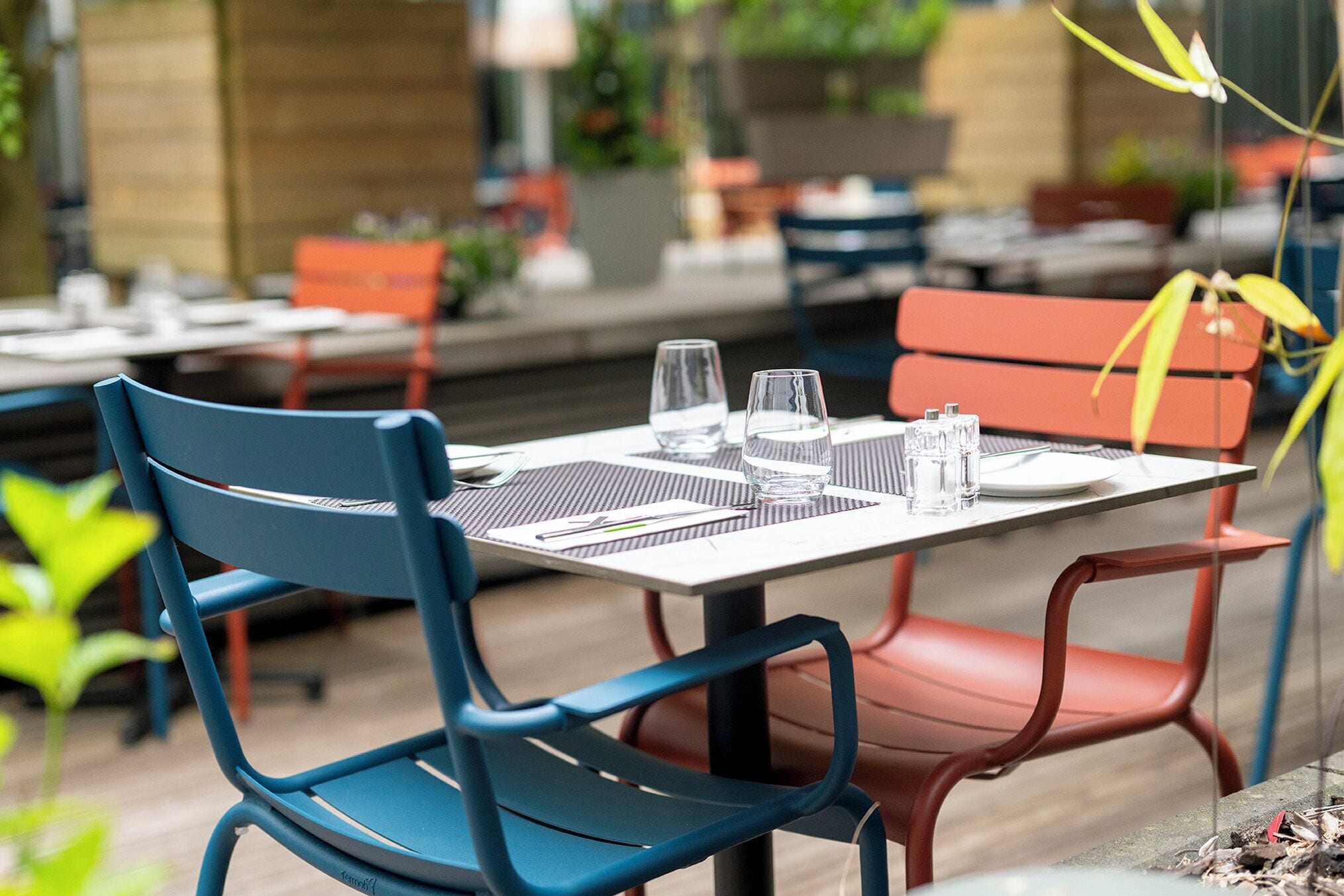 Comedor con mesas dispuestas para dos, sillas metálicas y una combinación de asientos rojos y azules.