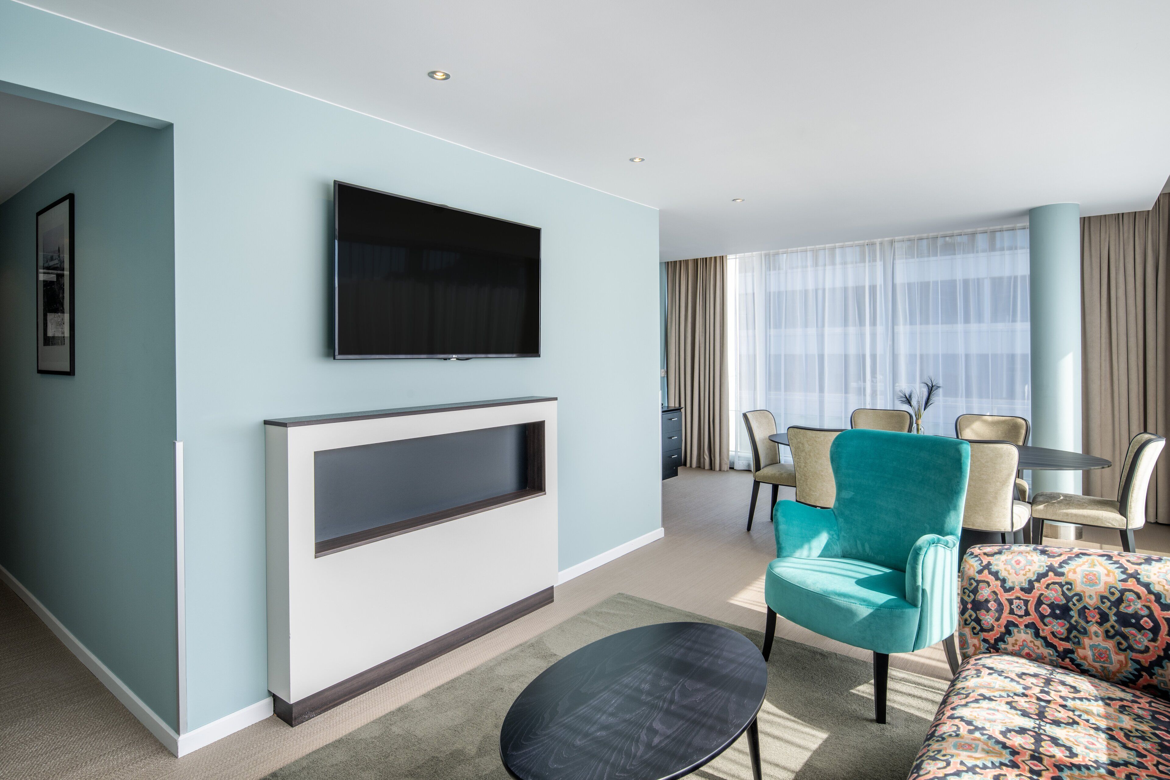 Una habitación de hotel moderna con televisión de pantalla plana montada en la pared, un sofá, una mesa de centro y un área de comedor con una mesa y sillas.