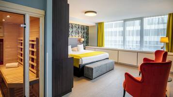 A hotel room with a bed, a bench, a red armchair, and a large window with curtains.