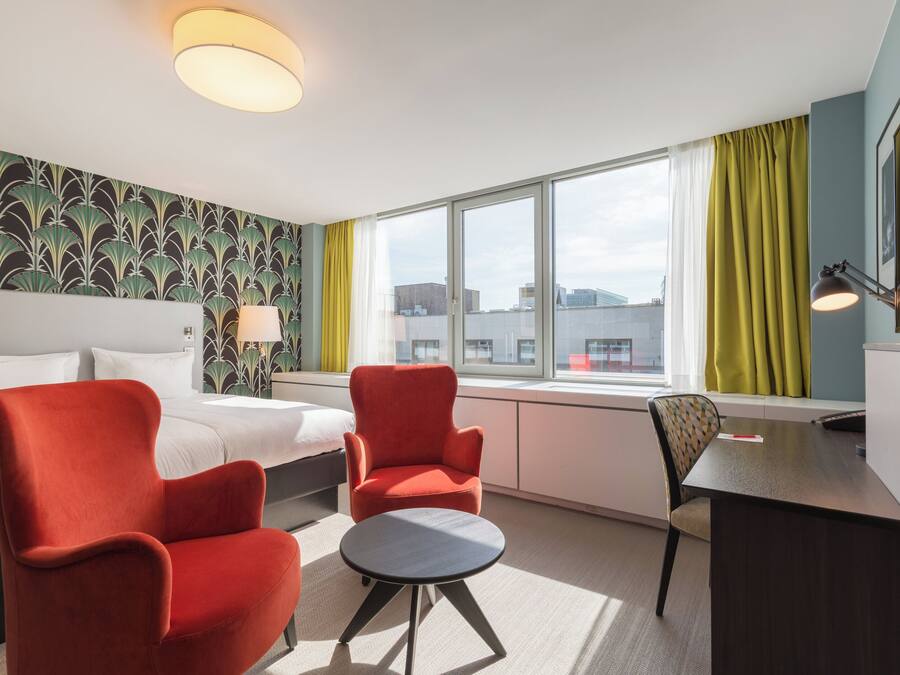 A hotel room with a bed, two red armchairs, a small round table, a desk with a chair, a window with curtains, and a wall with a leafy pattern.