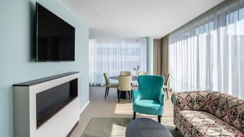 Un saló modern amb un televisor de pantalla plana muntat a la paret, un butac armchair color esmeralda, un sofà amb dibuixos i una zona de menjador amb taula i caires al fons.