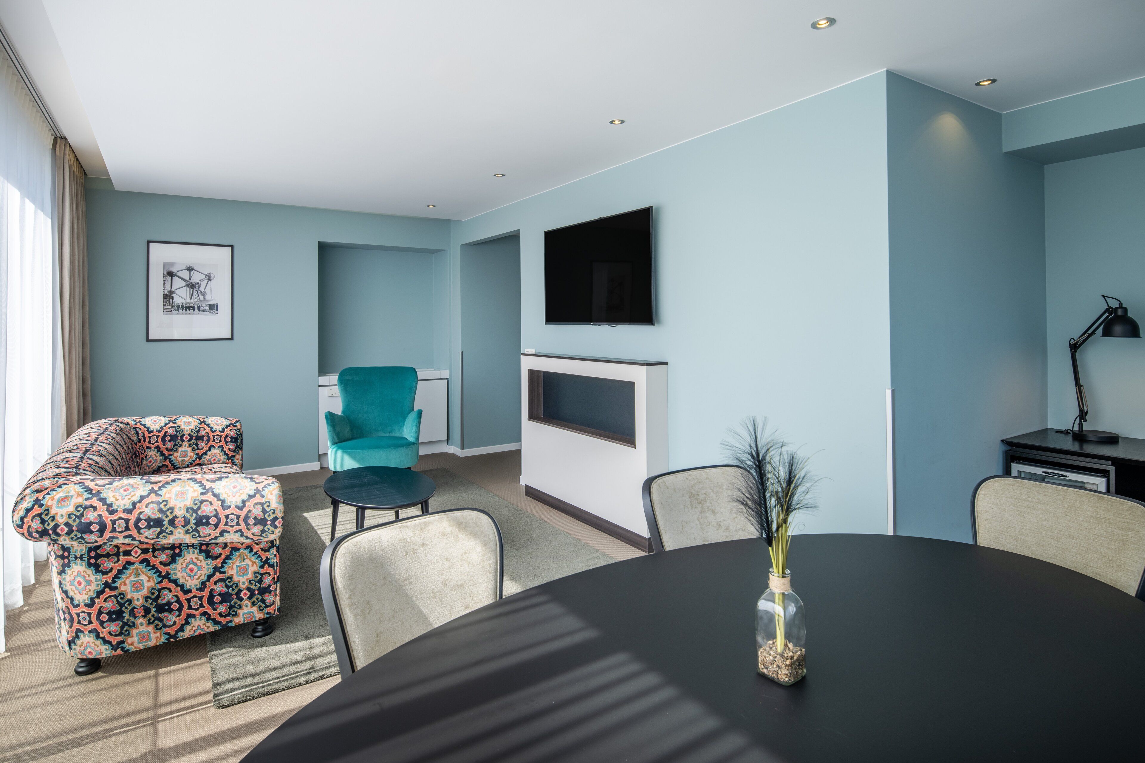 Een moderne woonkamer met een bank, een teal fauteuil en een eettafel met een vaas.
