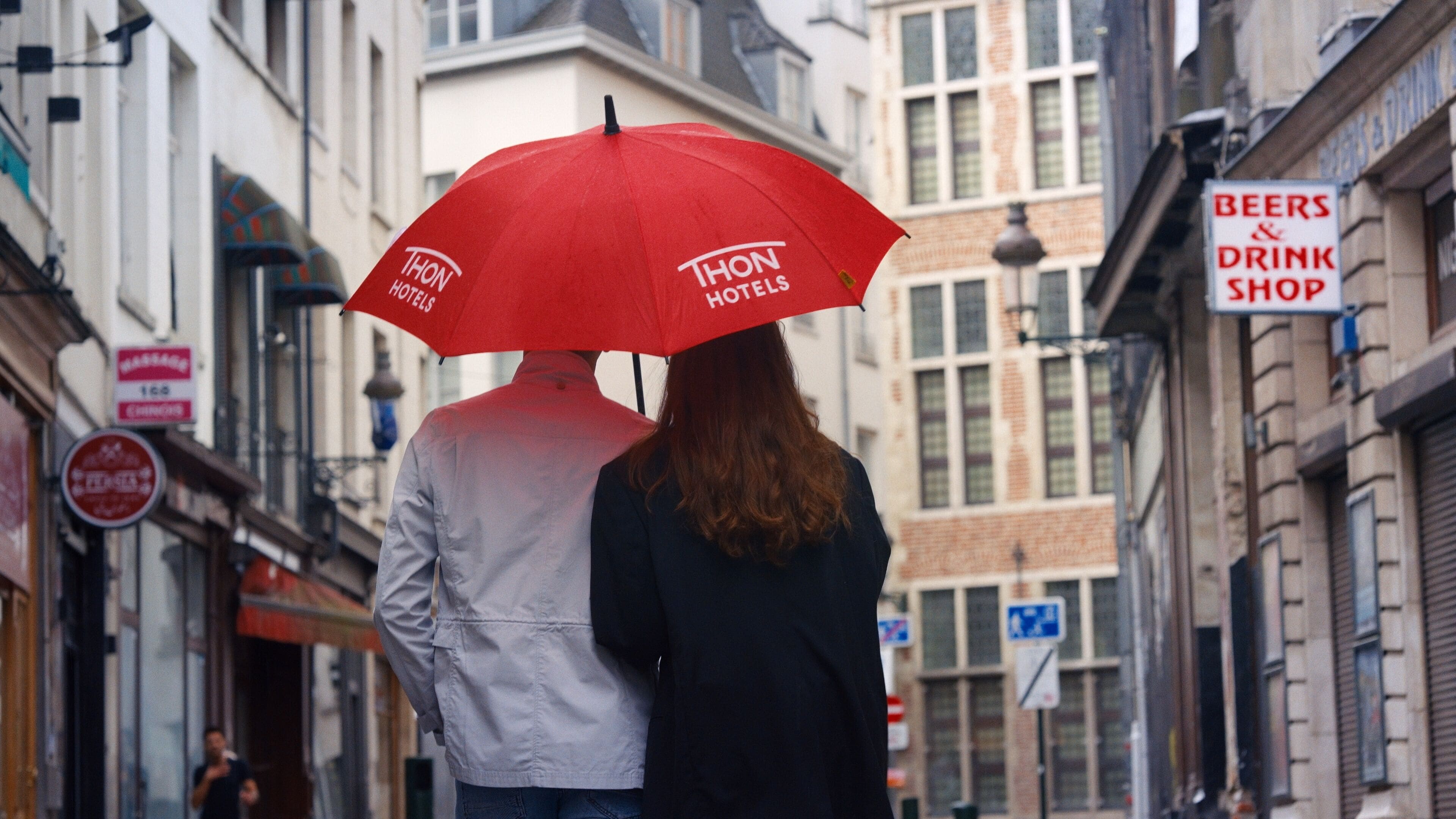 Um casal caminhando pela rua com um guarda-chuva vermelho, passando por um hotel e uma loja com placas.