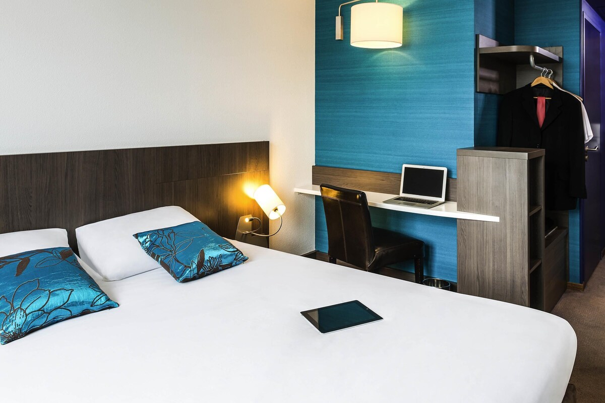 A hotel room with a bed, a desk with a laptop, a chair, and a wardrobe.