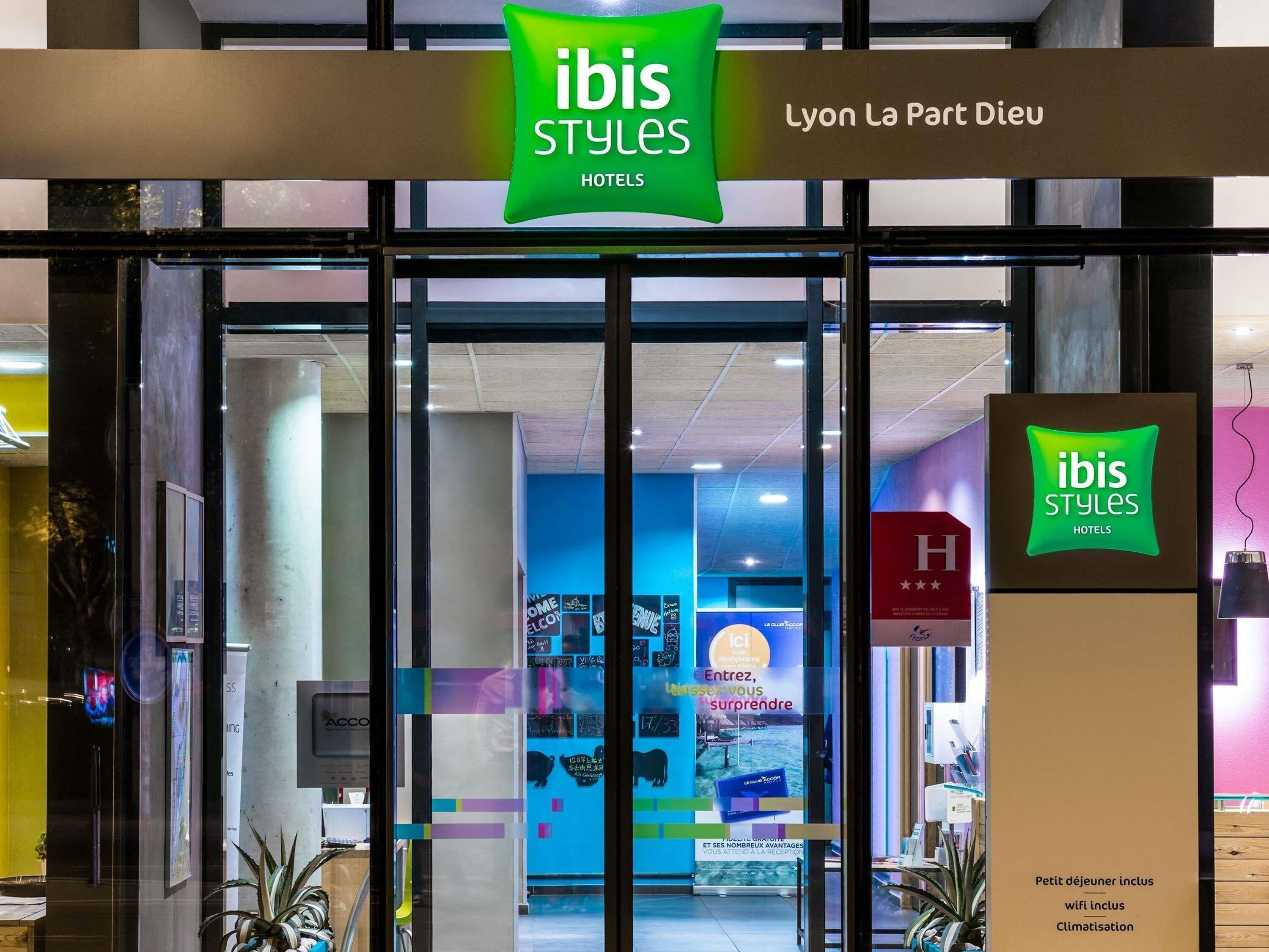The entrance to an ibis Styles hotel with a glass door, signage, and a visible interior with a blue wall and promotional posters.