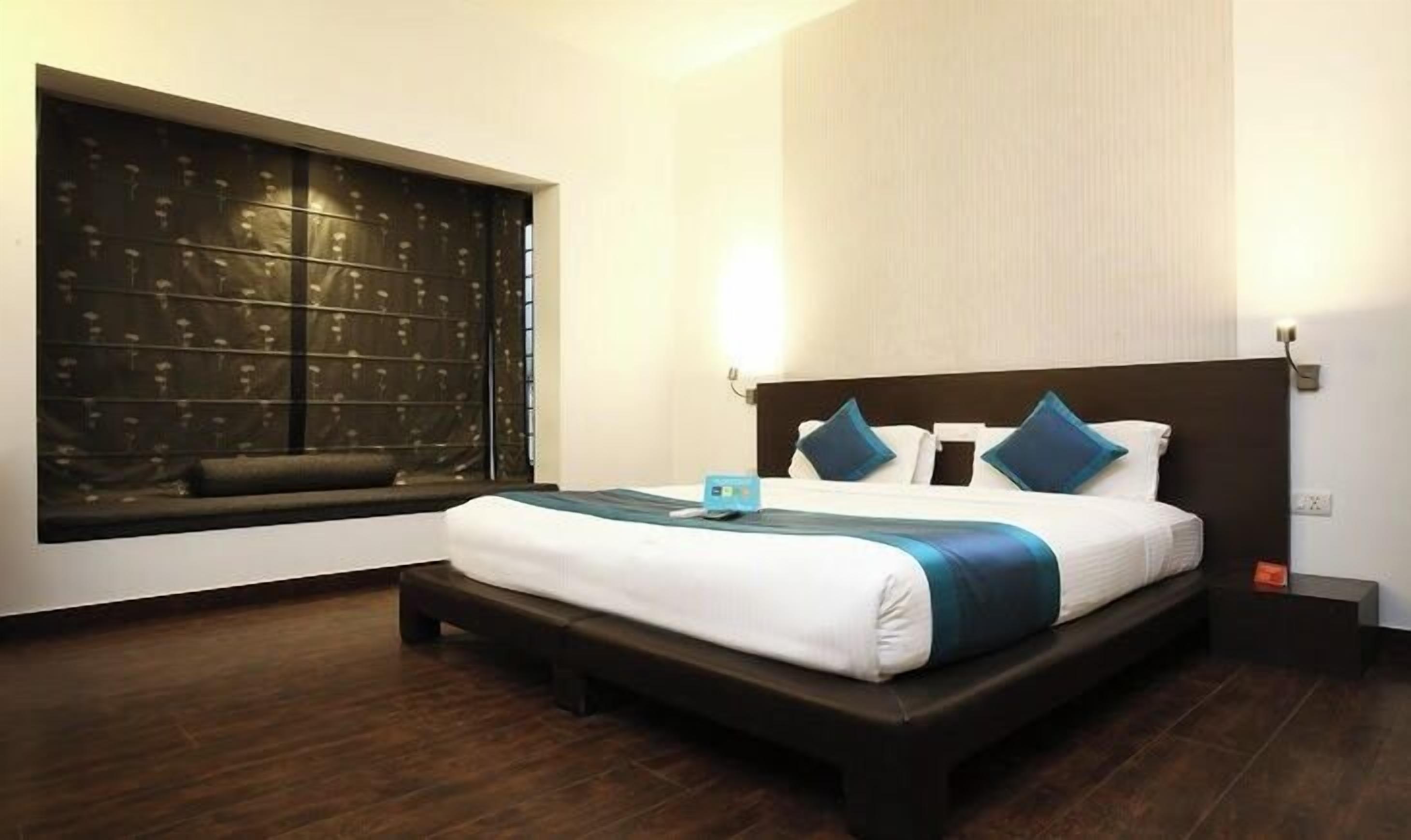 Una habitación de hotel con una cama grande, una ventana con persianas, una mesita de noche y una lámpara fijada en la pared.