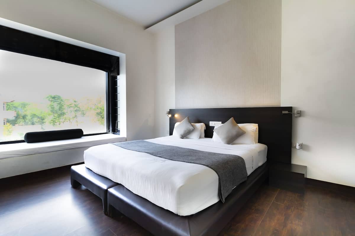 A modern bedroom with a large bed, a bench at the foot of the bed, and a window with a view of greenery.
