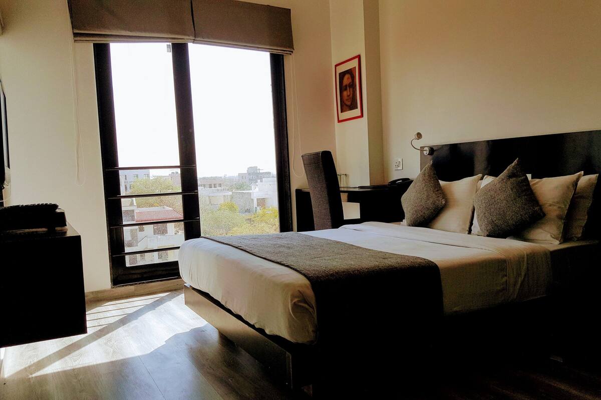 A hotel room with a large bed, a desk, a chair, and a window with a city view.