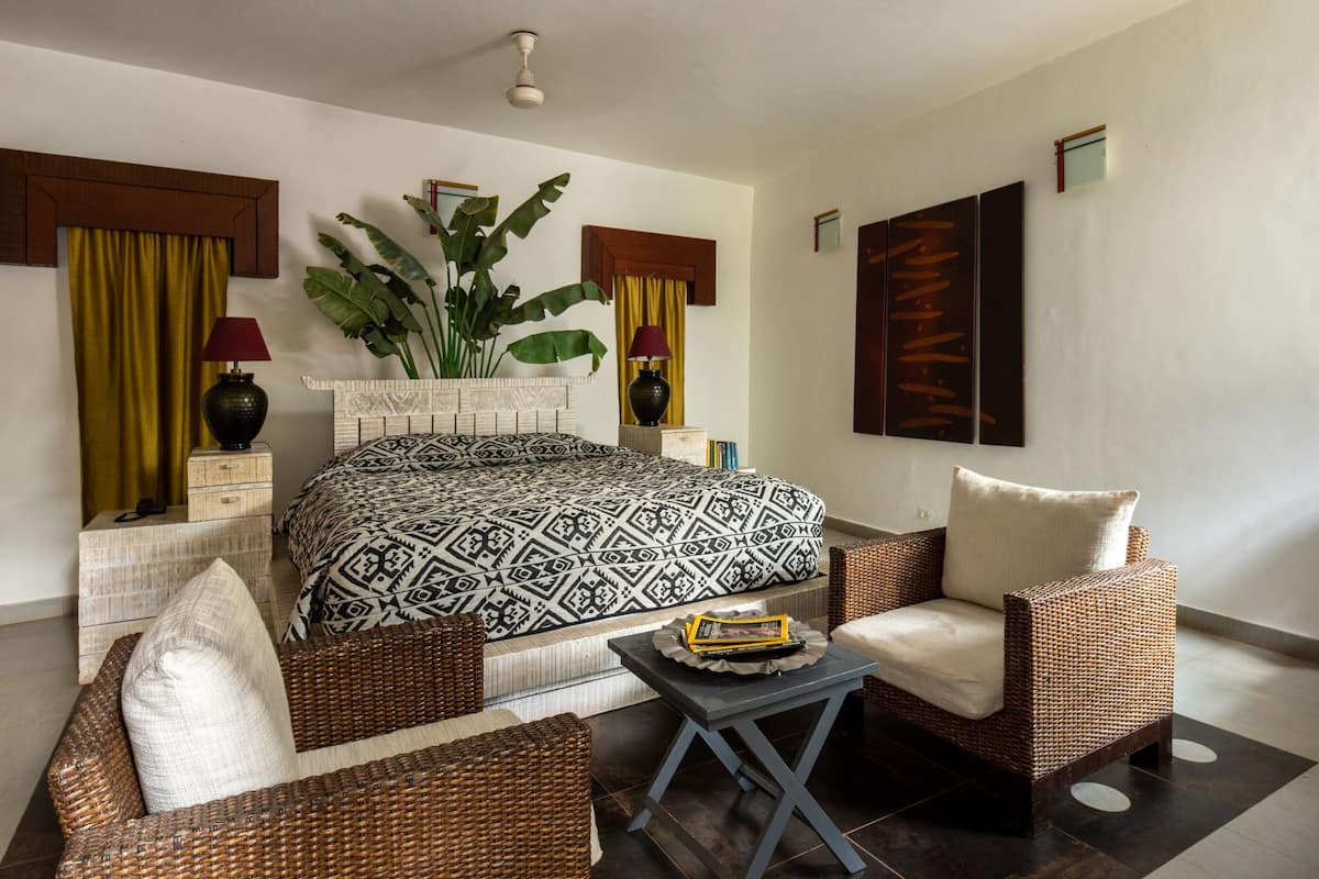 A hotel room with a large bed, two wicker chairs, a small table, a potted plant, and a painting on the wall.