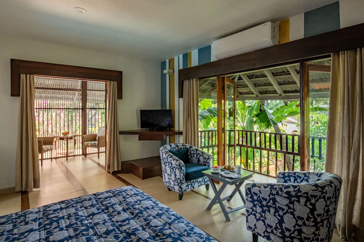 A room with a bed, two patterned chairs, a small table, a TV, and a balcony with a view of greenery.