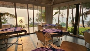 A room with a view of the ocean, featuring a table and chairs with checkered tablecloths, a ceiling fan, and large windows. - Casa Vagator (Vagator)