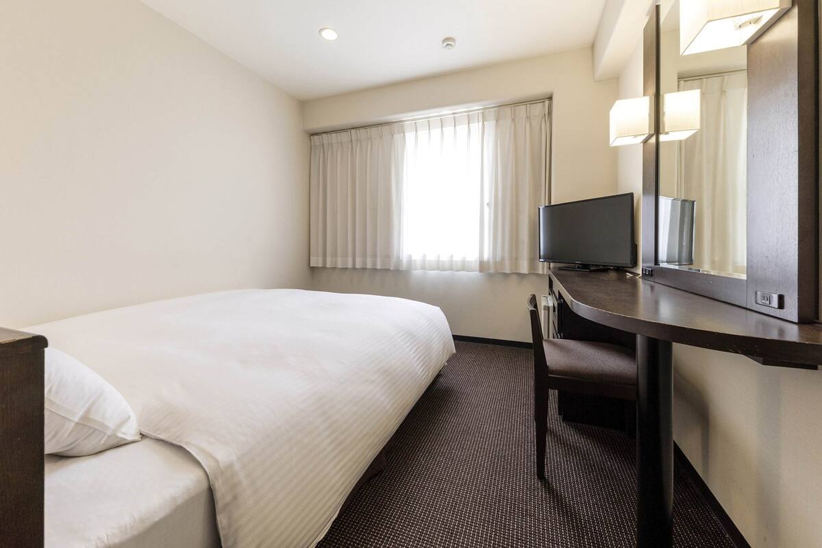 A hotel room with a bed, a desk with a flat-screen TV, a mirror, and a window with curtains.