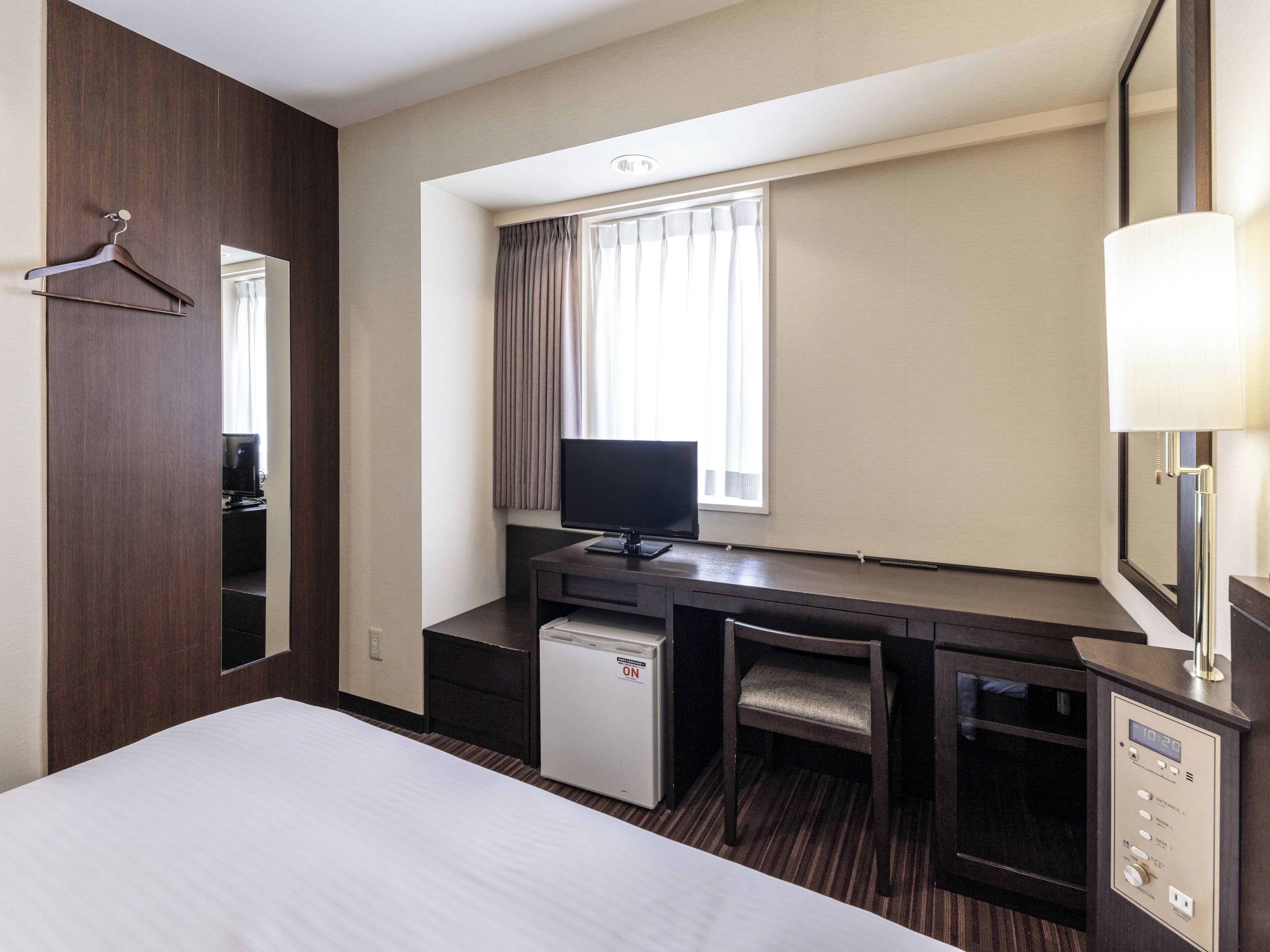 A hotel room with a bed, a desk with a computer, a TV, a mini-fridge, a chair, and a mirror.