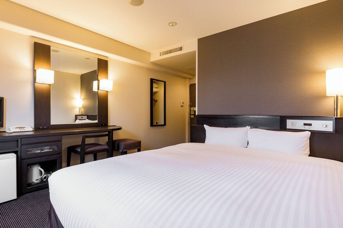 A hotel room with a bed, a desk with a television, a mirror, and a bedside table with a lamp.