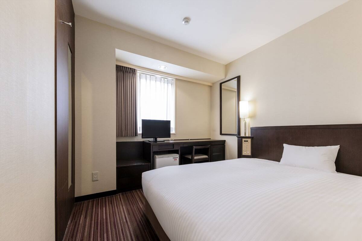 A hotel room with a large bed, a desk with a computer, a television, a mirror, and a window with curtains.