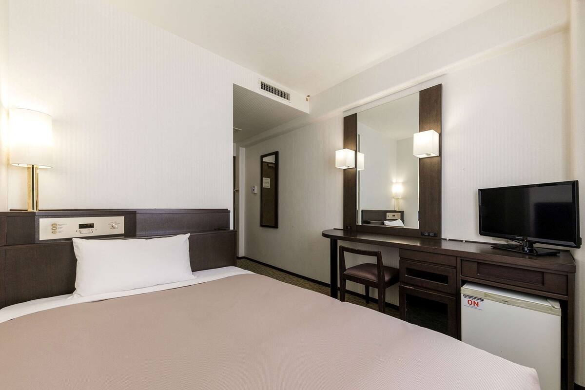 A hotel room with a bed, a desk with a television, a chair, and a small refrigerator.
