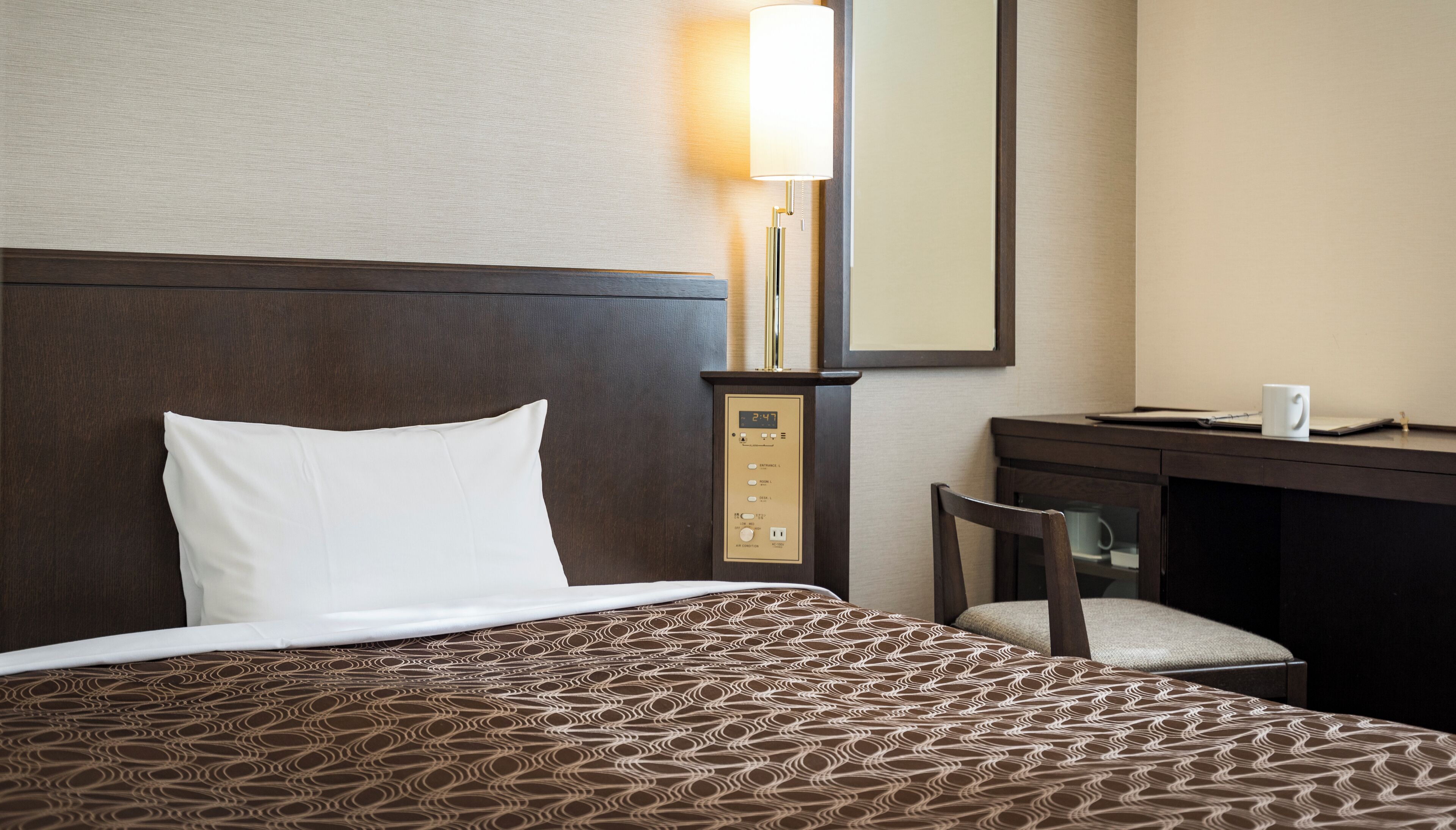 A hotel room with a bed, a desk, a chair, a lamp, and a control panel.