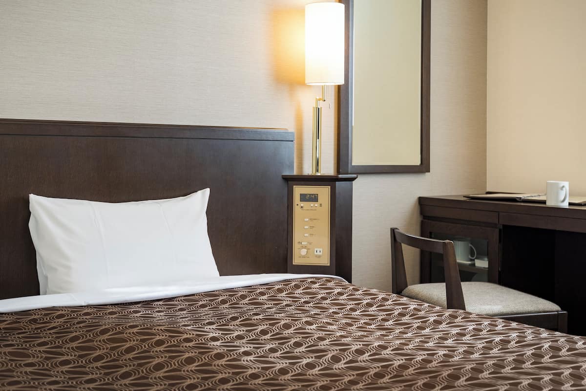 A hotel room with a bed, a desk, a chair, a lamp, and a control panel.