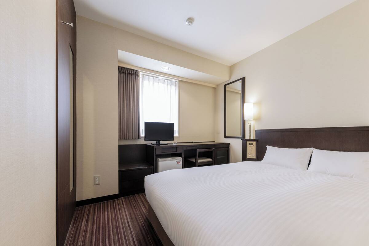 A hotel room with a large bed, a desk with a computer, a chair, a TV, and a mirror.