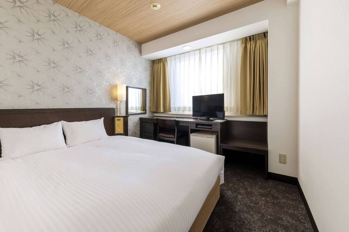 A hotel room with a large bed, a desk with a TV, a desk lamp, and a window with curtains.