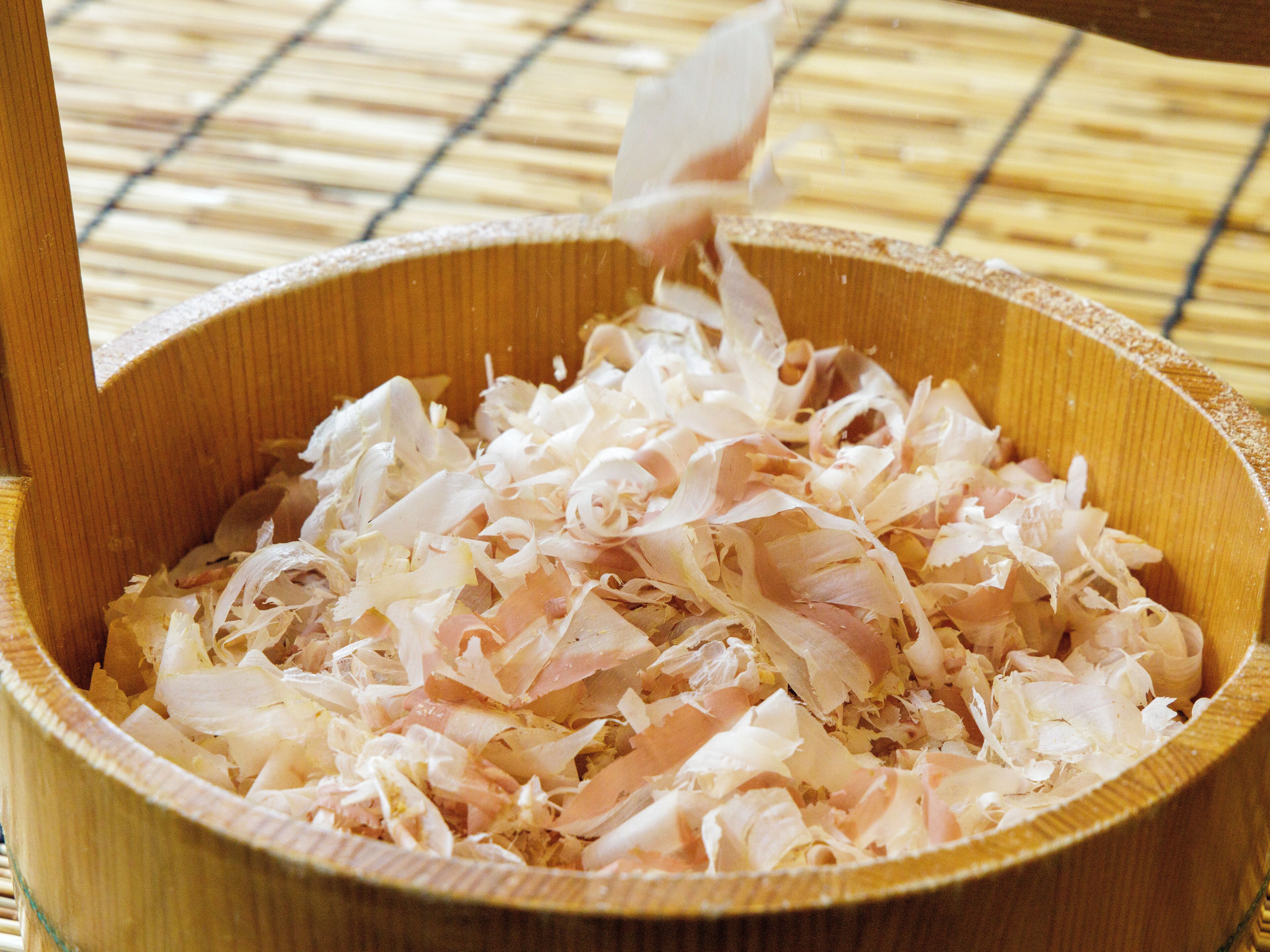 A wooden bowl filled with shredded white material, possibly food, on a bamboo mat.