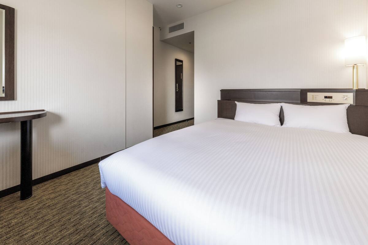 A hotel room with a large bed, a bedside table, a mirror, and a wall-mounted control panel.