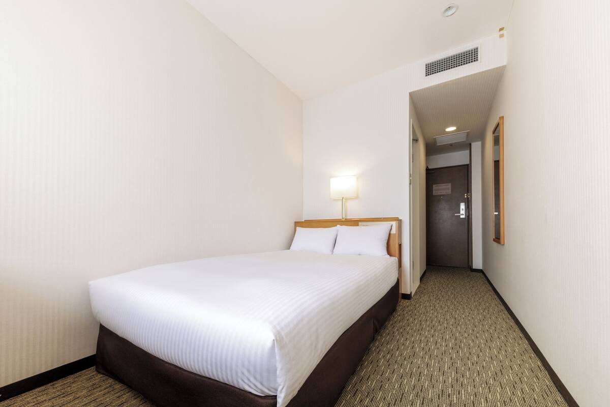 A hotel room with a single bed, a bedside lamp, and a carpeted floor.