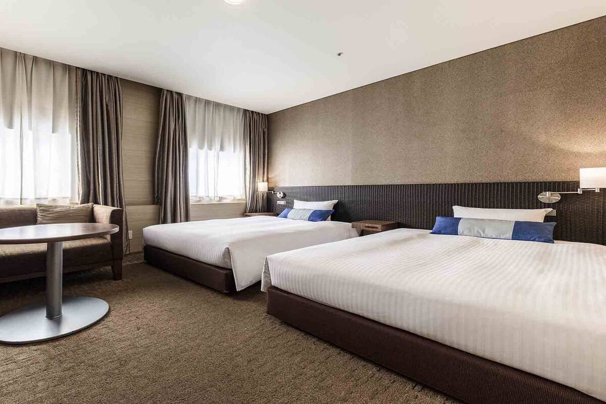 A hotel room with two beds, a sofa, a round table, and a large window with curtains.