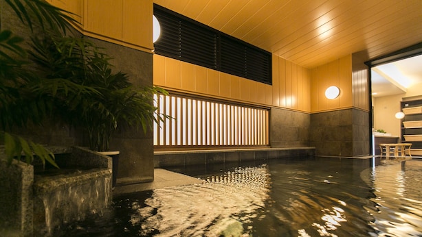 A serene indoor pool area with a stone water feature, wooden paneling, and a seating area with a round light fixture. - Nishitetsu Inn Kurosaki (Kitakyushu)