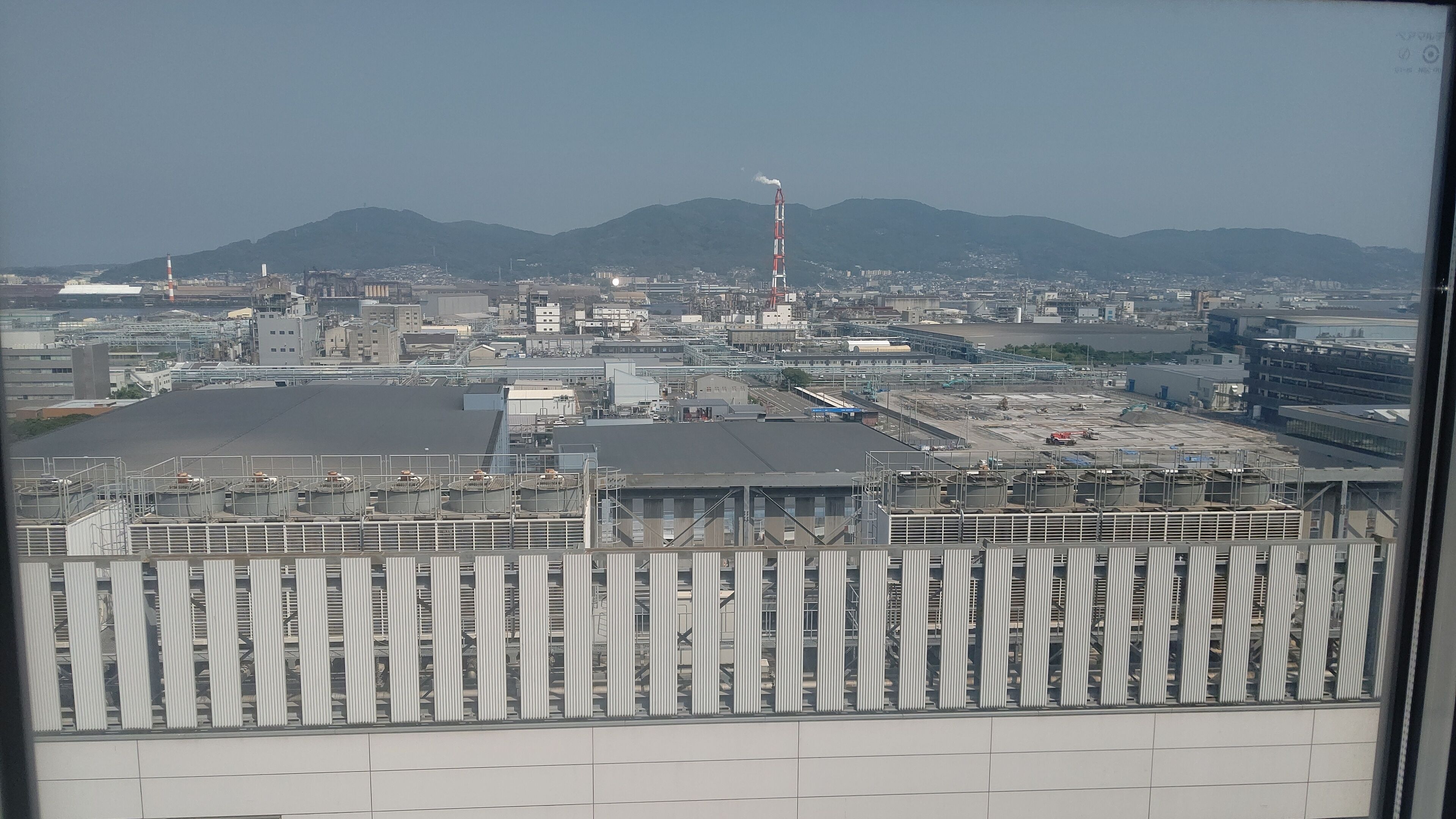 A large industrial facility with multiple buildings, a prominent smokestack, and a mountainous backdrop.