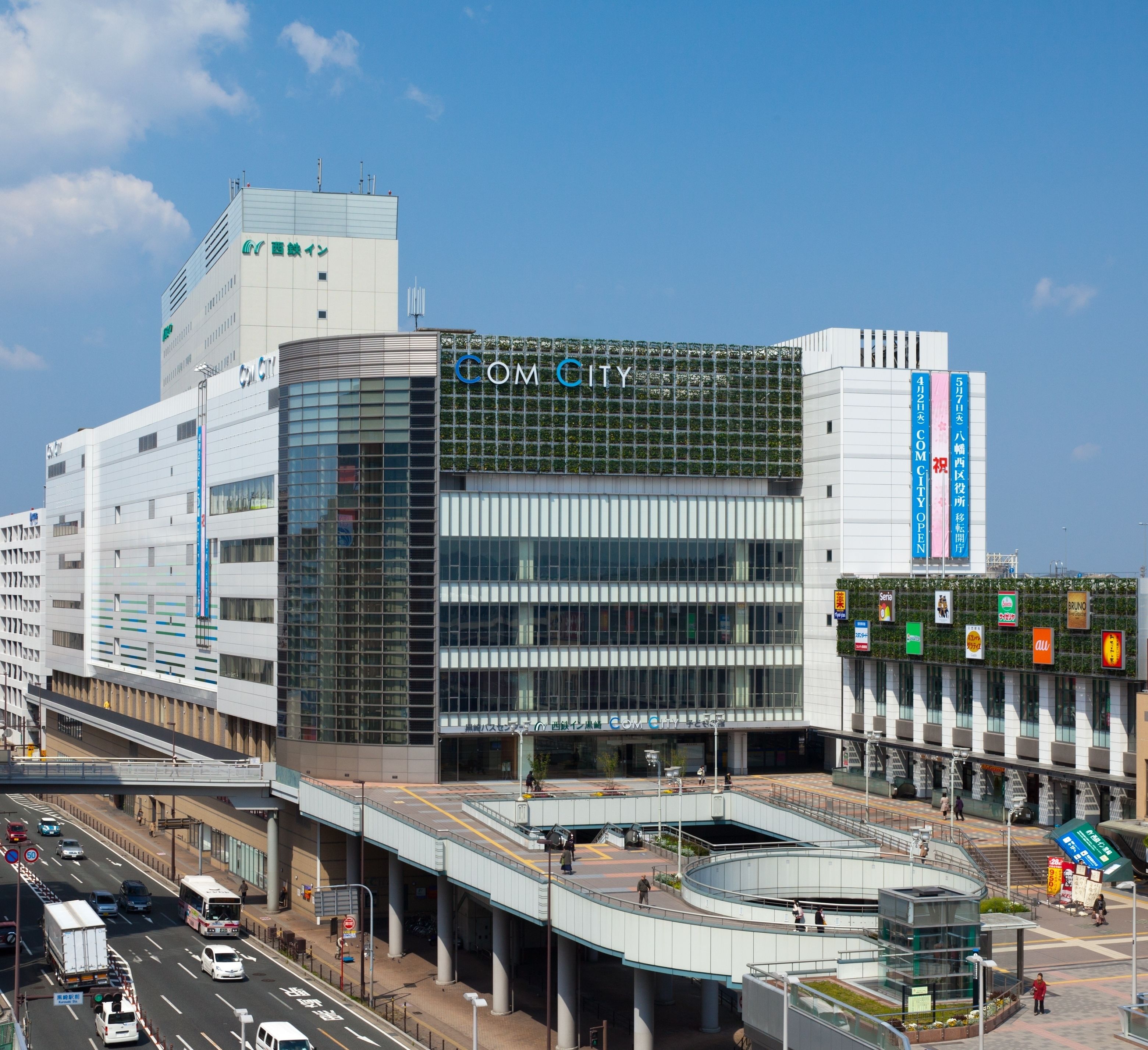 A modern building with a prominent "COM CITY" sign, featuring a glass facade and a covered pedestrian area.