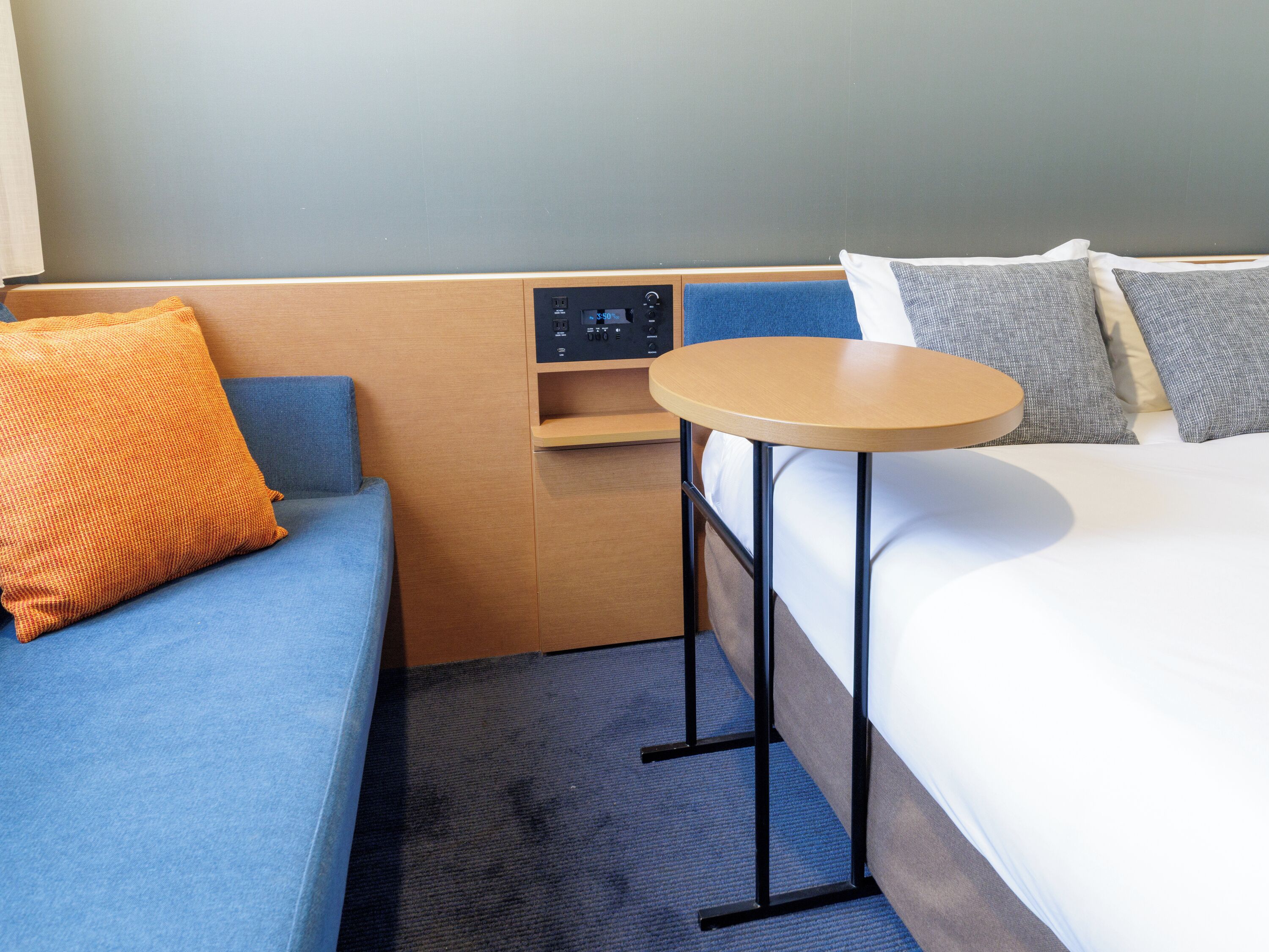 A hotel room with a bed, a small table, a sofa, and a control panel.