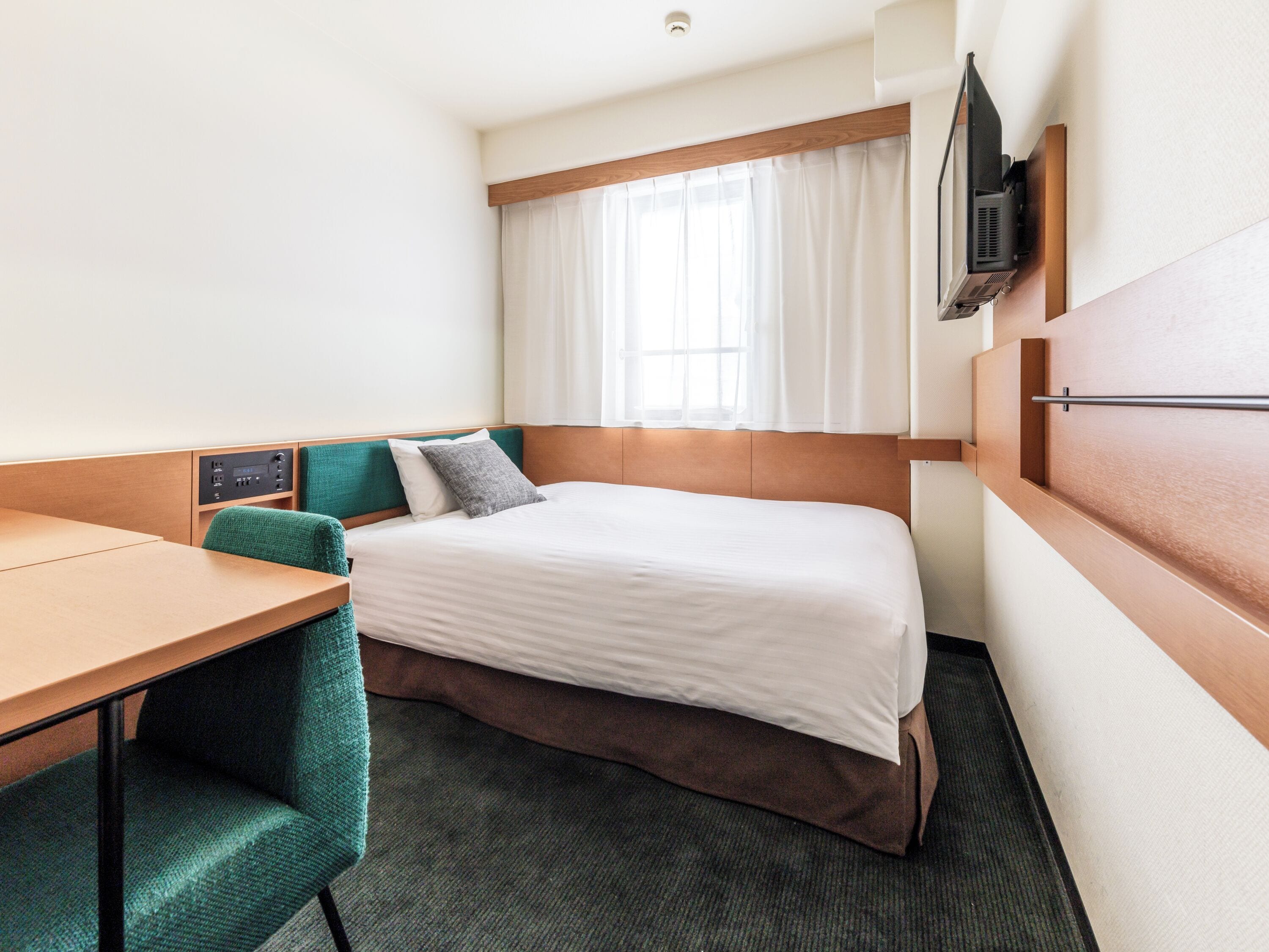 A hotel room with a bed, a desk, a chair, a television mounted on the wall, and a window with curtains.