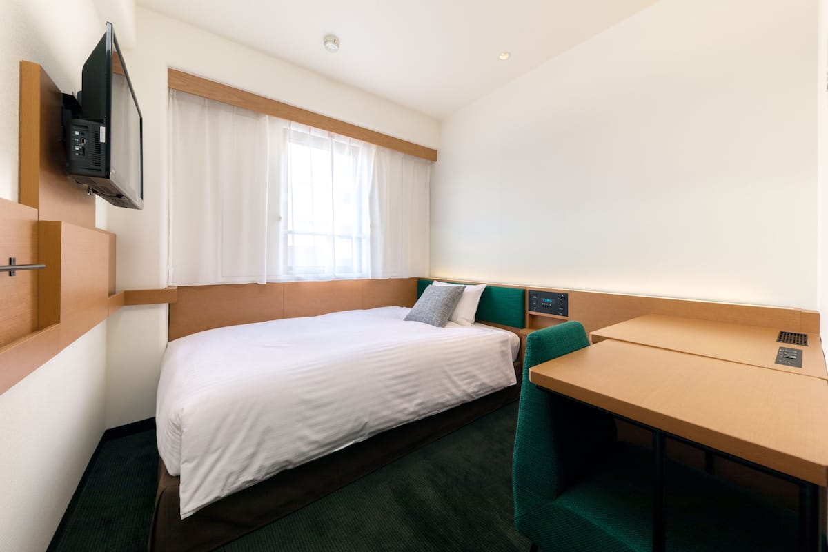 A compact hotel room with a single bed, a desk, a chair, a TV mounted on the wall, and a window with curtains.