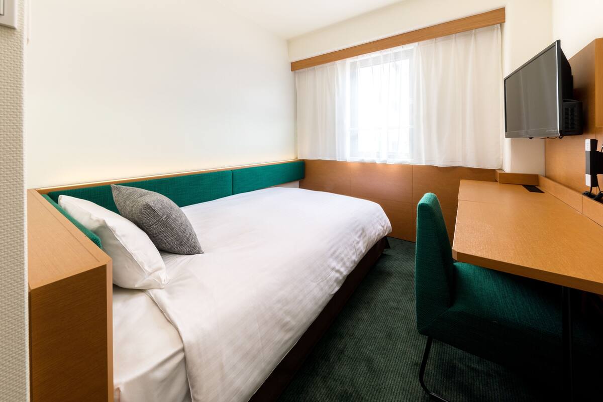A hotel room with a single bed, a desk, a chair, a television, and a window with curtains.