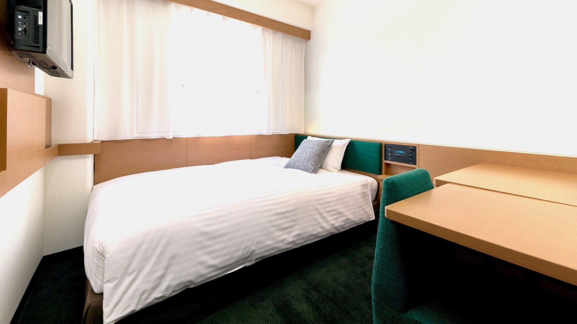 Un lit simple avec un drap blanc, une chaise verte, une petite table et un téléviseur fixé au mur.