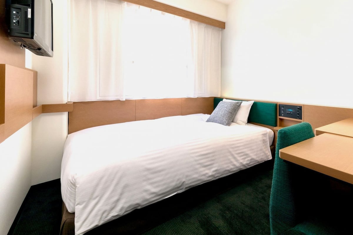A single bed with a white sheet, a green chair, a small table, and a TV mounted on the wall.