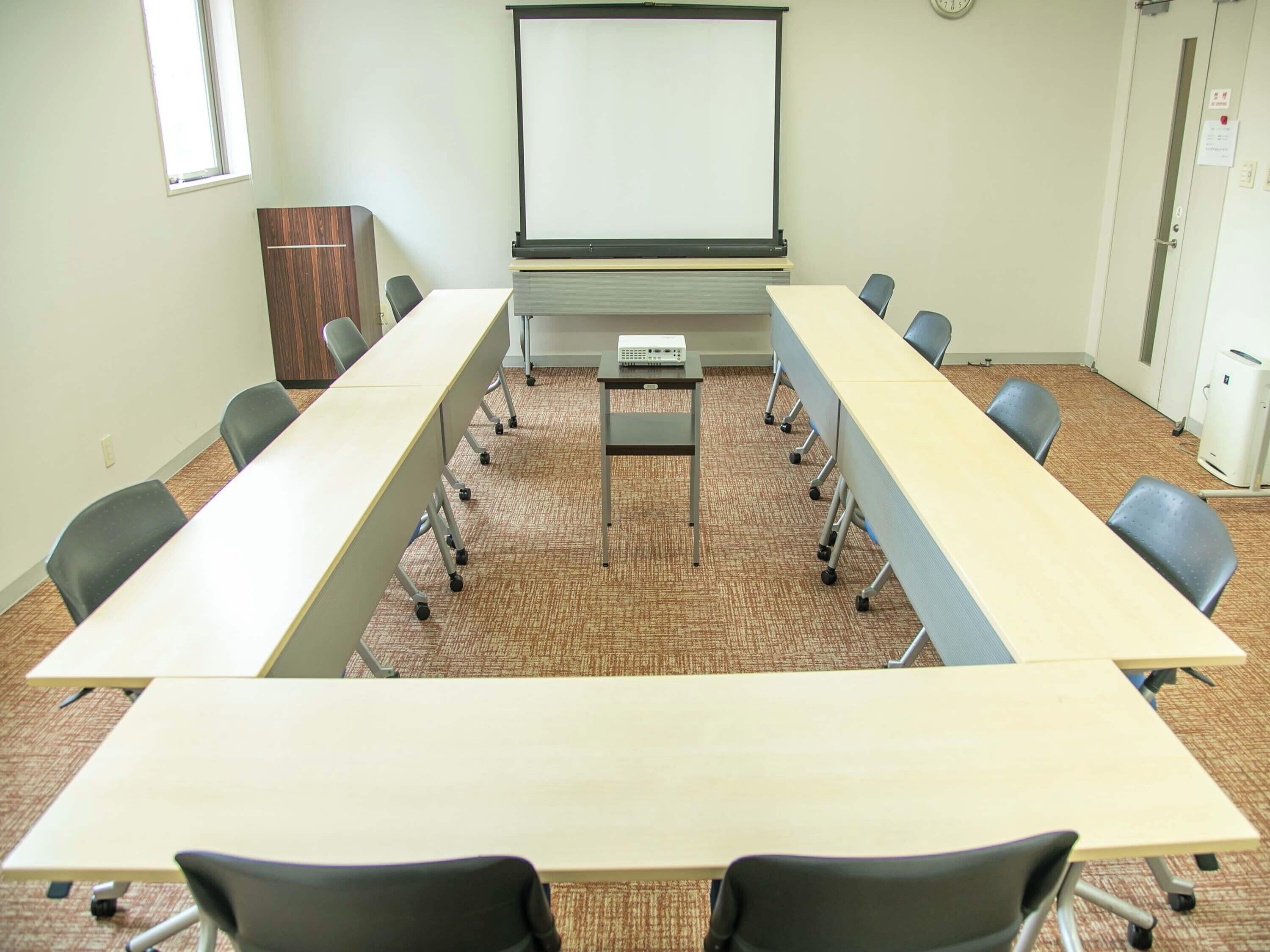 Un salón de conferencias con una larga mesa, sillas, una pantalla de proyector y un reloj en la pared.