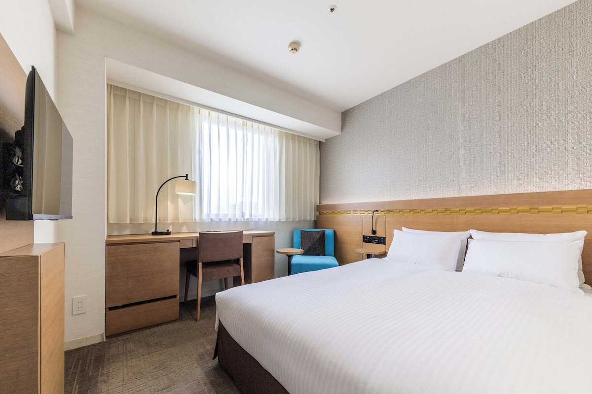 Une chambre d’hôtel avec un lit, un bureau, une chaise, une télévision et une fenêtre avec des rideaux.