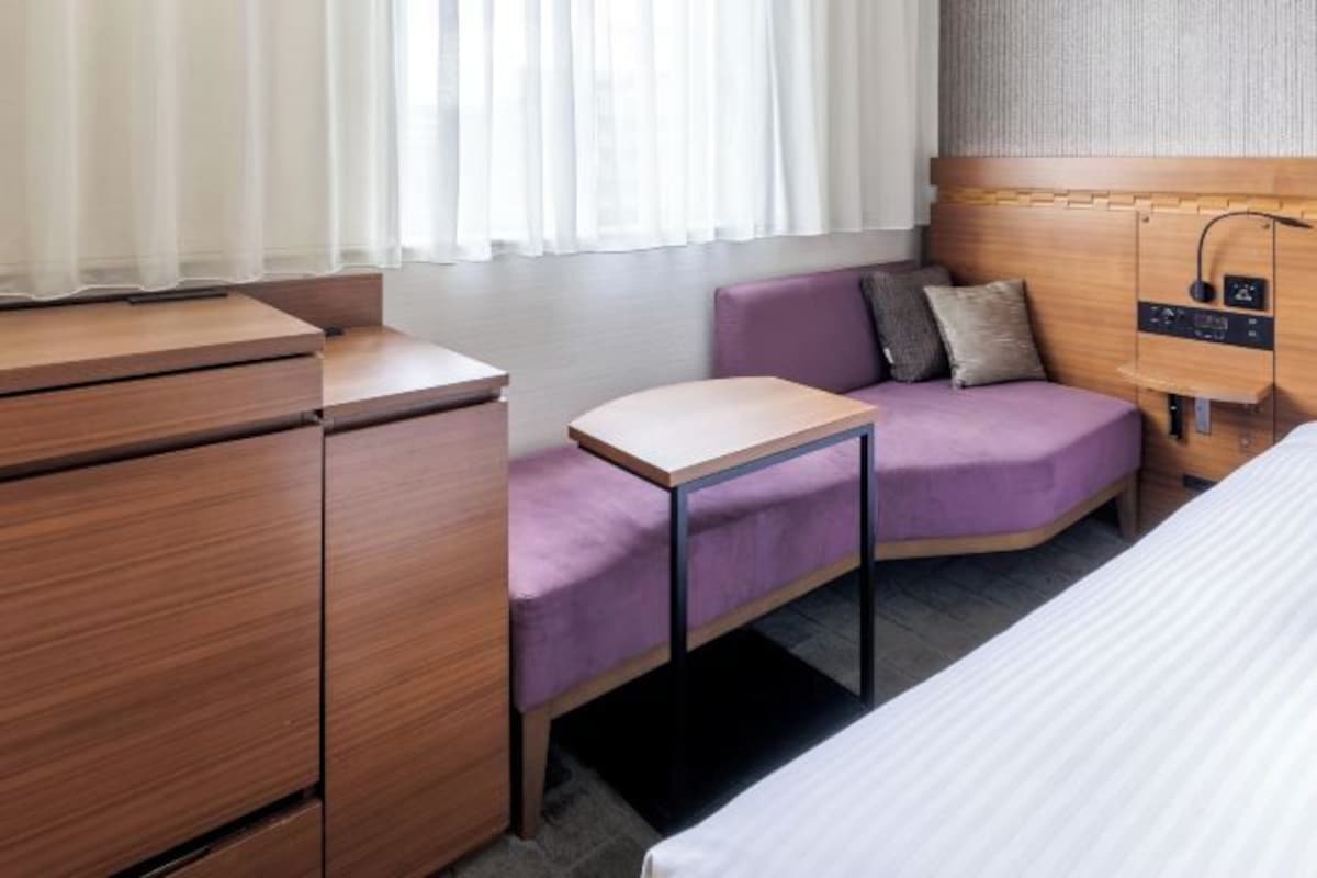 Een compacte hotelkamer met een bed, een klein bureau, een zithoek met kussens en een nachtkastje.