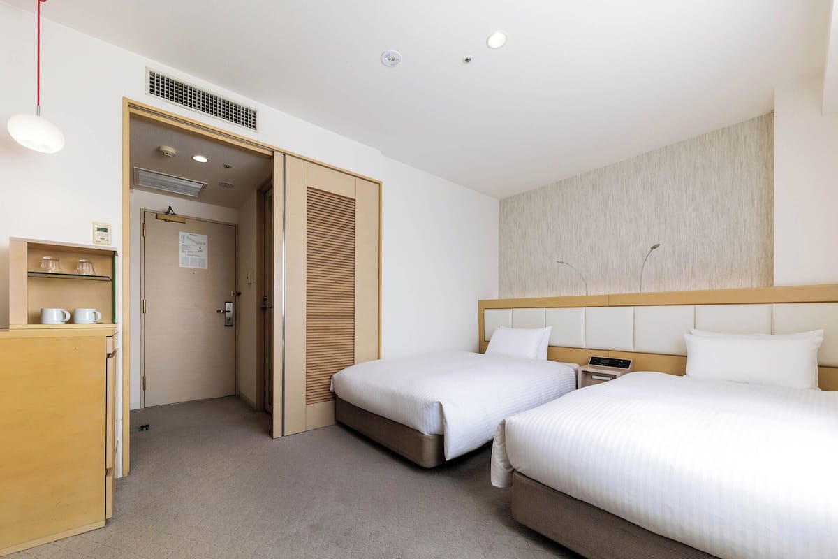 Une chambre d’hôtel avec deux lits, une armoire en bois contenant des tasses, une porte avec une grille d’aération et un luminaire.