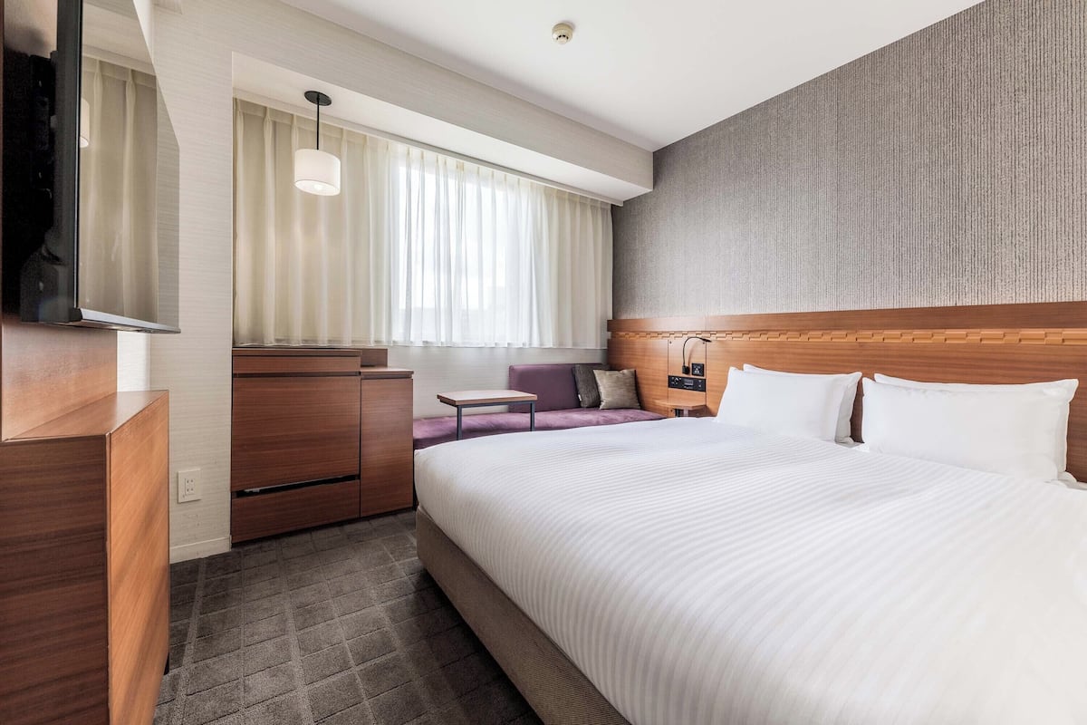 Une chambre d’hôtel moderne avec un grand lit, une tête de lit en bois, un bureau avec une chaise, une petite table et un téléviseur fixé au mur.