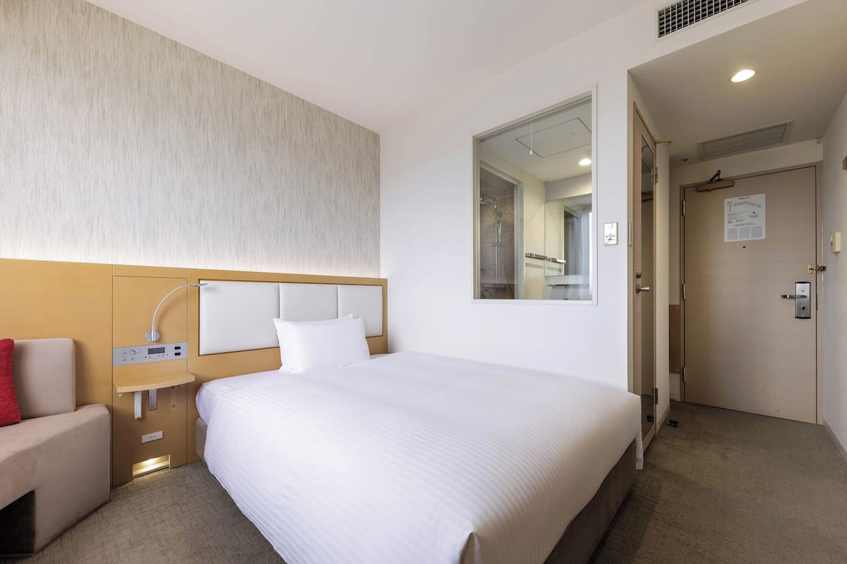Une chambre d’hôtel avec un grand lit, une table de chevet avec une lampe, un petit coin salon et une douche visible à travers une porte en verre.