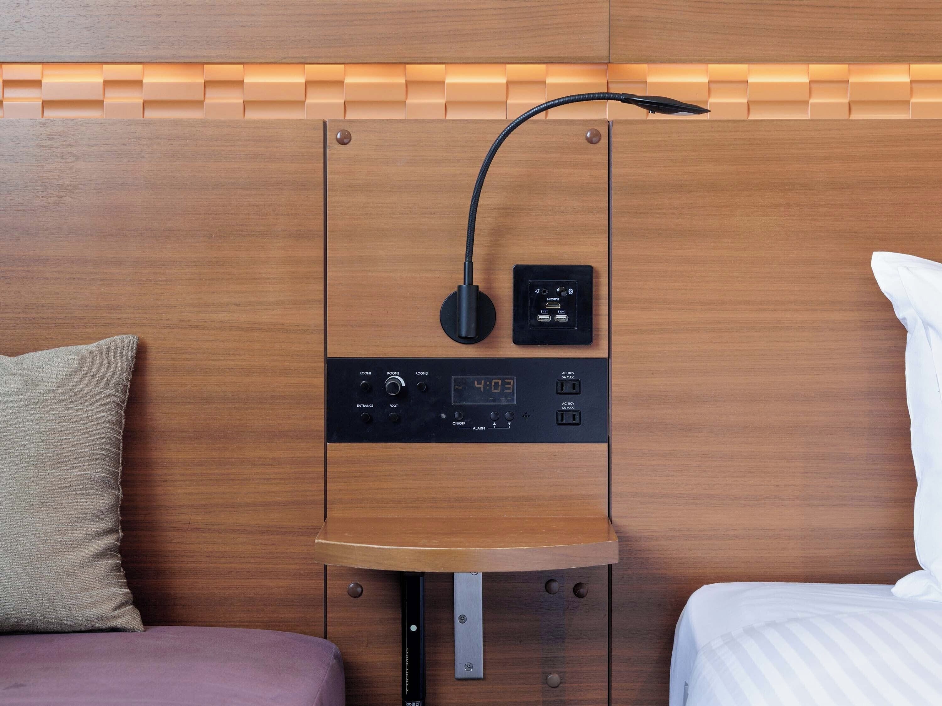 A modern hotel room with a wooden headboard, a control panel with a digital clock, and a small table with a lamp.