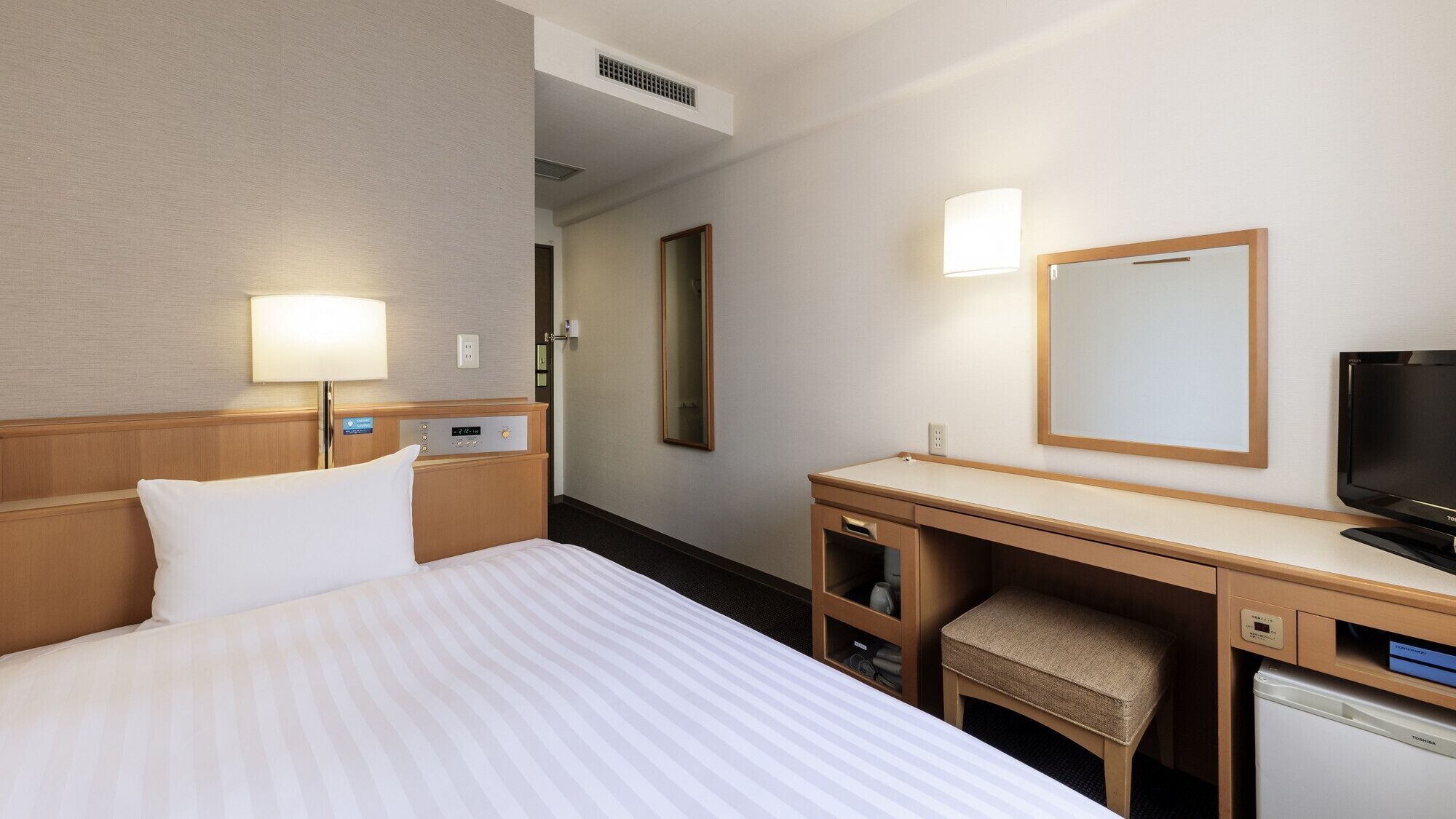 A hotel room with a bed, bedside table, lamp, TV, desk, chair, and a mirror.