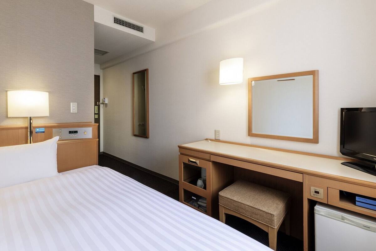 A hotel room with a bed, bedside table, lamp, TV, desk, chair, and a mirror.